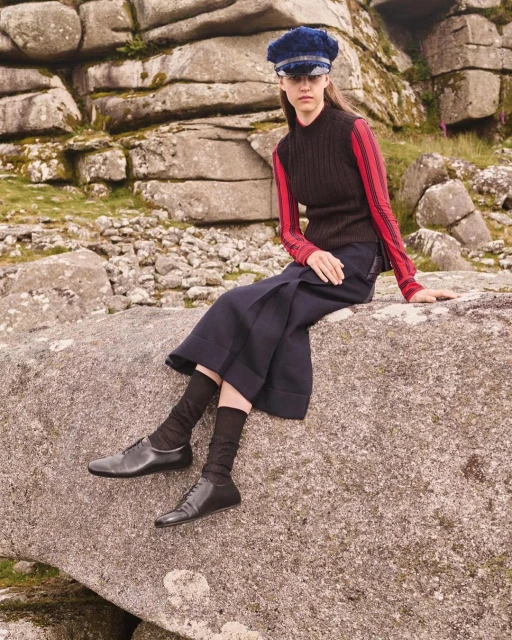 A young woman in a dark knit top, wide pants, and a cap sits on a large rock with a rocky landscape background in a fashion editorial.