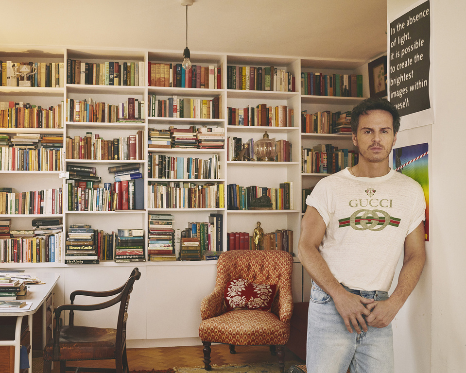 Man wearing a white graphic t-shirt and light blue jeans standing in a room with a large white bookcase filled with books