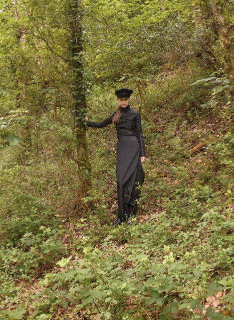 A woman in a black outfit stands in a lush green forest, leaning against a tree. The natural light filters through the dense foliage.