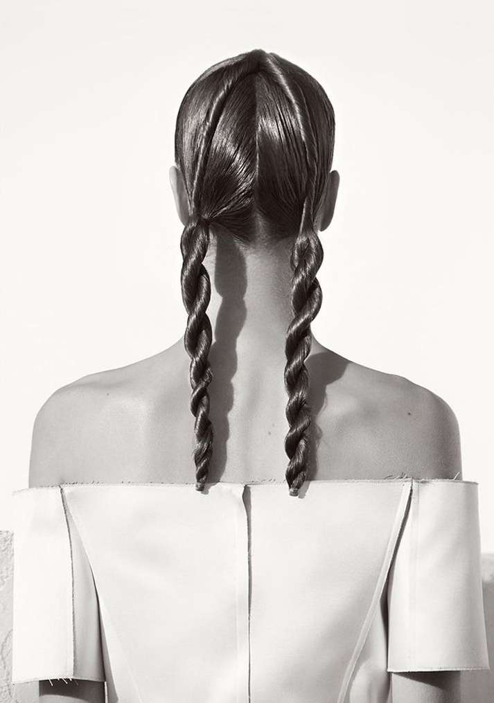 Back view of a woman with long brown hair styled in two spiral braids, wearing a light-colored, off-the-shoulder top against a white background.