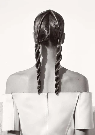 Back view of a woman with long brown hair styled in two spiral braids, wearing a light-colored, off-the-shoulder top against a white background.