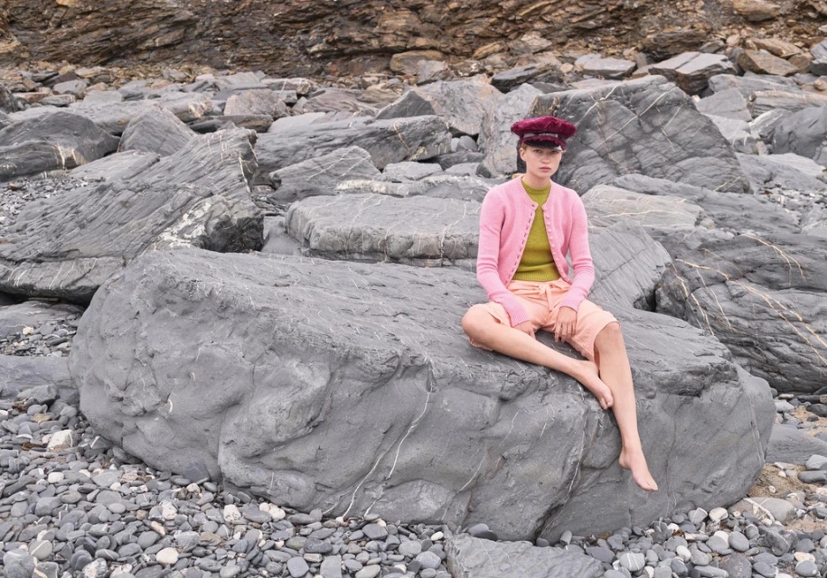 A young fair-skinned woman in casual pink cardigan and hat sits barefoot on a large grey boulder at a rocky beach coastline.