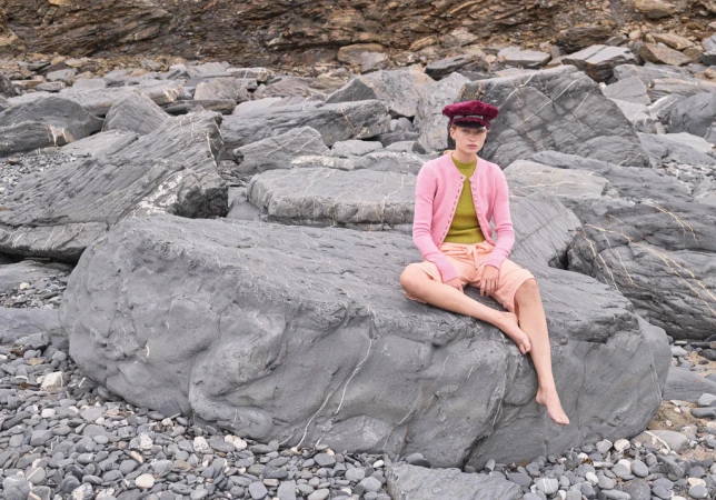 A young fair-skinned woman in casual pink cardigan and hat sits barefoot on a large grey boulder at a rocky beach coastline.