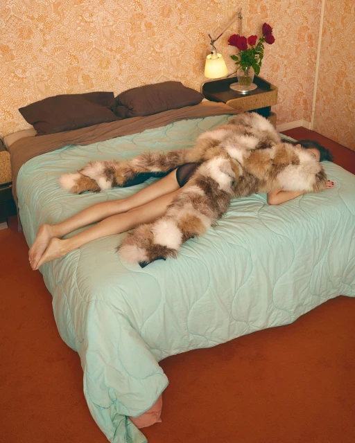 A young adult female is lying on a bed with a fur throw, soft lighting, and floral wallpaper in an indoor setting.