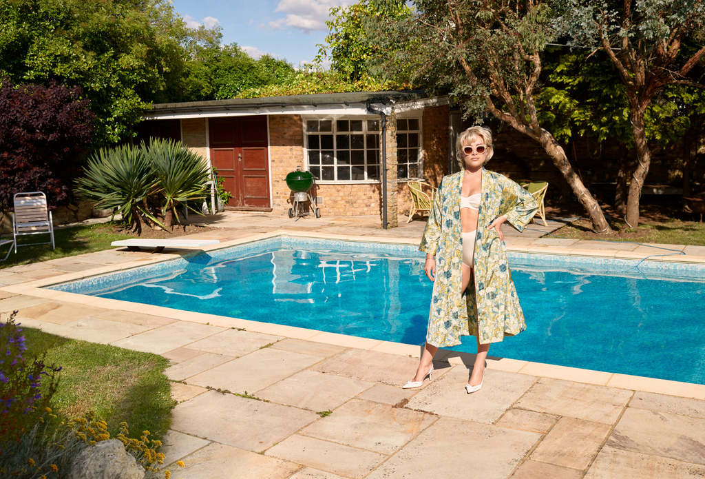 Adult woman in robe and bikini poses poolside in backyard with trees and building. Sunny, eye-level shot with natural light.