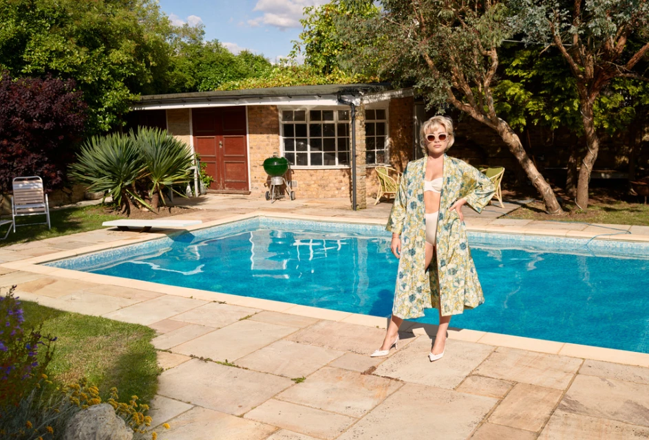 Adult woman in robe and bikini poses poolside in backyard with trees and building. Sunny, eye-level shot with natural light.