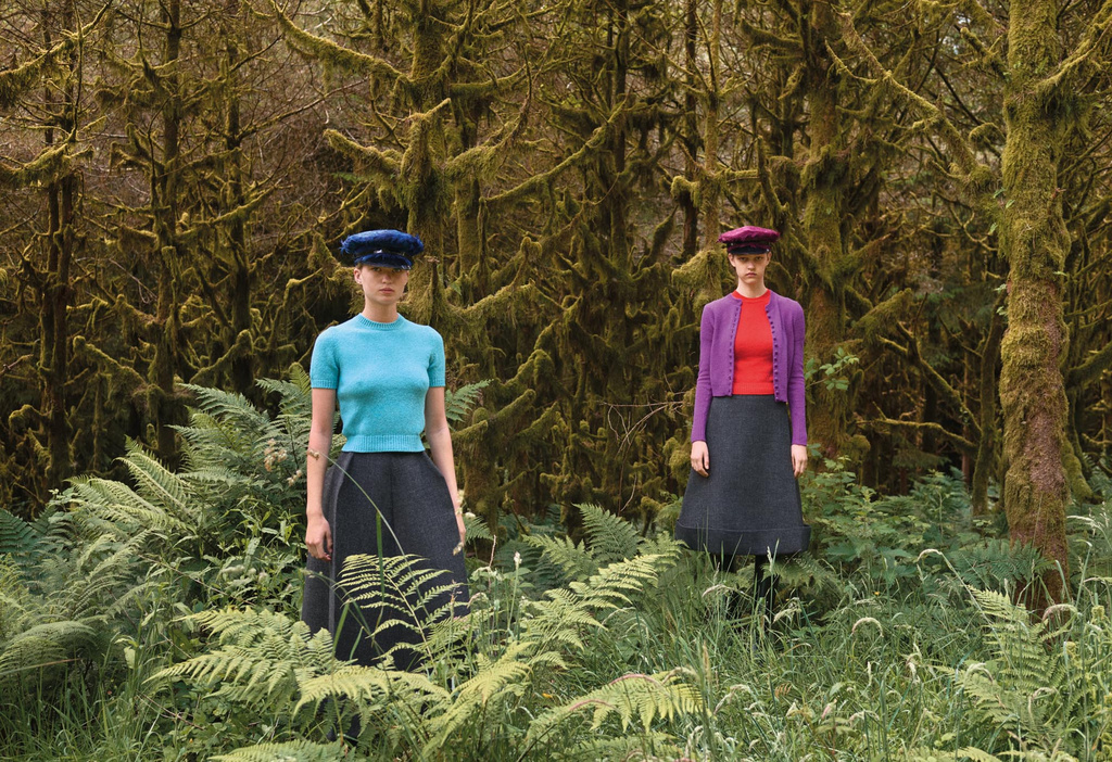 Two adult women wearing colored tops, dark skirts, and hats posing in a lush green forest with tall moss-covered trees in the background.