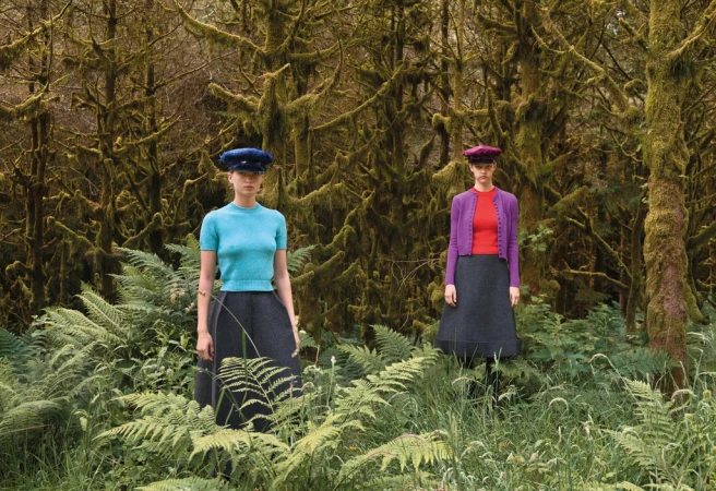 Two adult women wearing colored tops, dark skirts, and hats posing in a lush green forest with tall moss-covered trees in the background.
