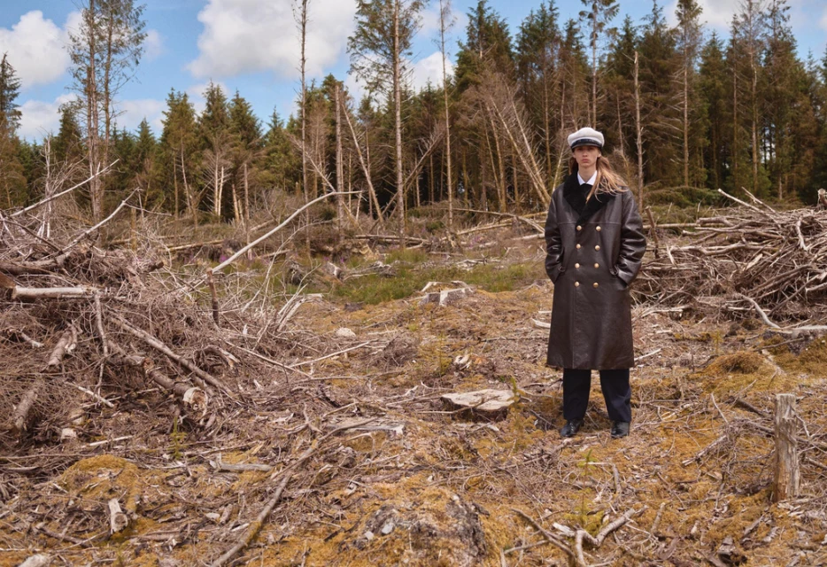 Woman in a coat standing in a logged forest area with fallen trees and green trees under a blue sky with clouds in an outdoor setting.