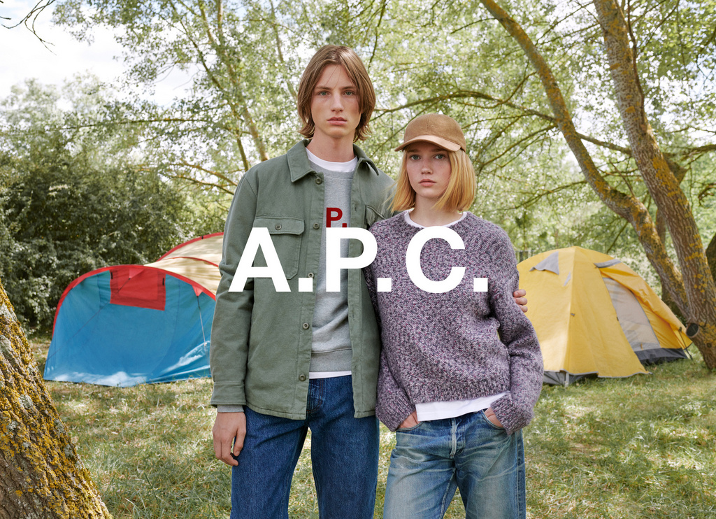 Fashion models pose outdoors in front of tents, wearing casual clothing. Image features neutral tones and natural lighting in a grassy area.