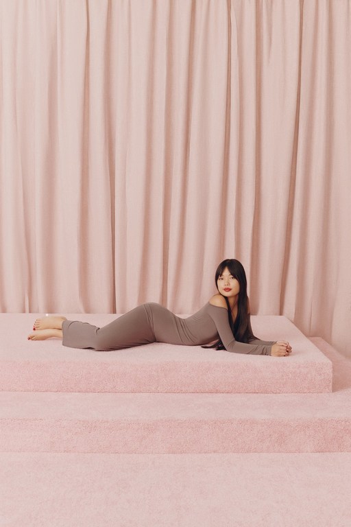 An Asian woman lying on pink steps, wearing a brown dress, in a studio setting with a pink curtain backdrop.
