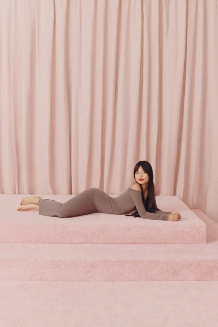 An Asian woman lying on pink steps, wearing a brown dress, in a studio setting with a pink curtain backdrop.