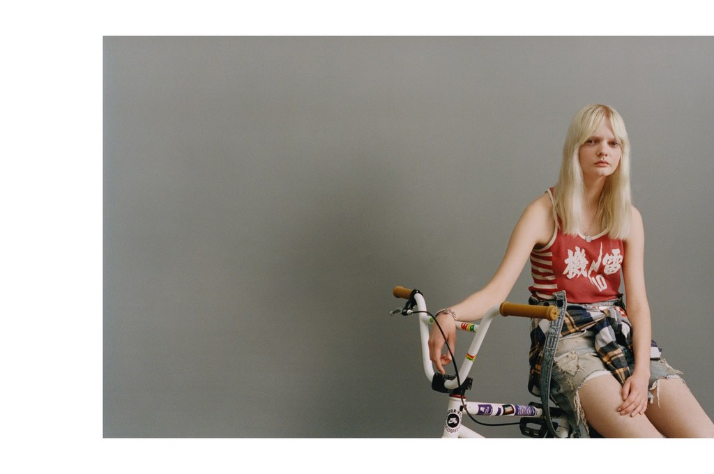 Young adult woman with blonde hair poses with a white BMX bike against a grey backdrop in a studio setting with soft, diffused lighting.
