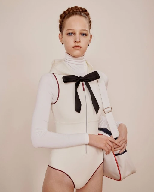 Young woman in white bodysuit and turtleneck poses in a studio with a sports bag. Braided hair, neutral makeup, and serious expression.