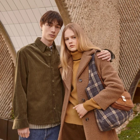 Two models posing outdoors with an olive corduroy shirt, brown wool coat, plaid shoulder bag, and structured leather handbag.