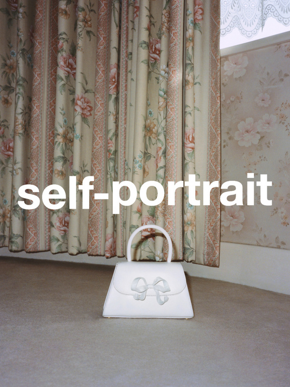 White purse with a bow sits on a neutral carpet in front of floral wallpaper and curtains in an indoor setting, close-up shot.