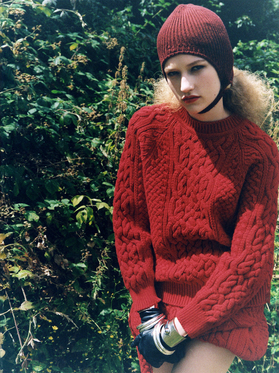 A young female model is posing outdoors in a red knit sweater, matching knit cap, black gloves, and metal arm cuffs in a natural setting.