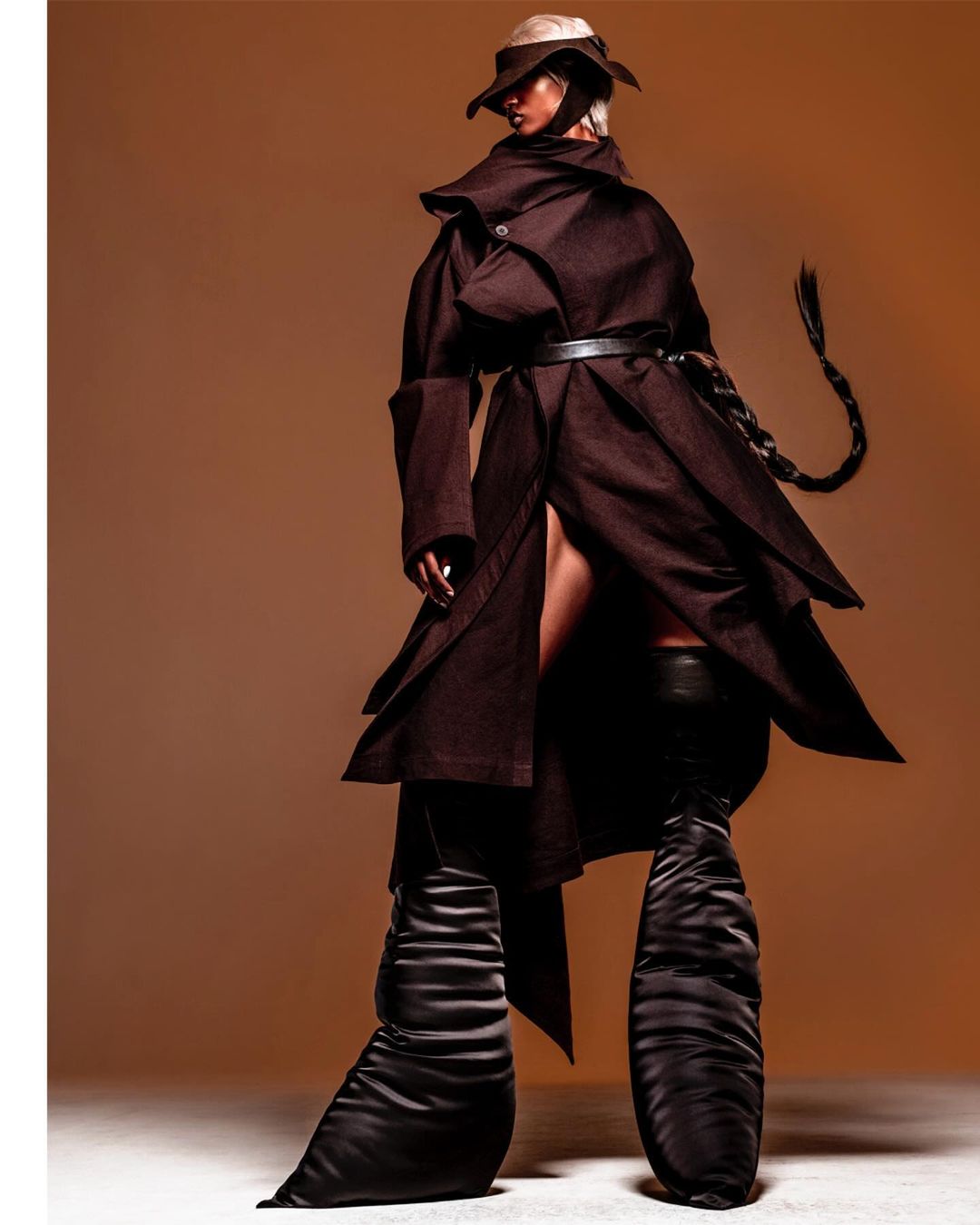 Fashion model poses in a brown belted coat and oversized black boots against a warm gradient background in a studio shot.