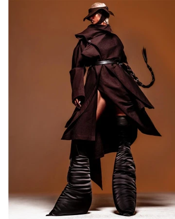 Fashion model poses in a brown belted coat and oversized black boots against a warm gradient background in a studio shot.