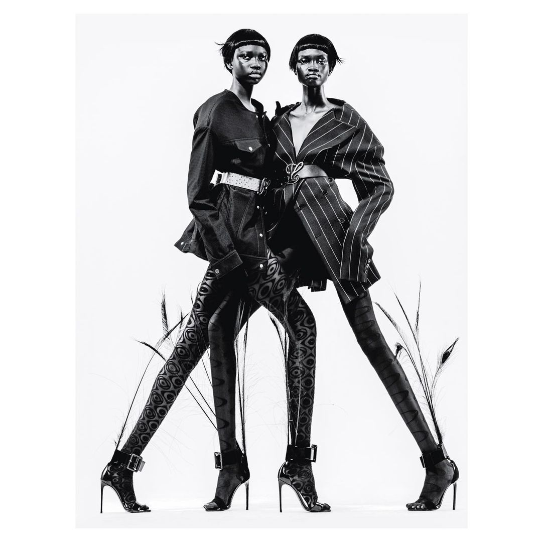 Two black women in fashionable black outfits and high heels pose in a studio, accented by stylized feather stalks in a black and white studio shot.