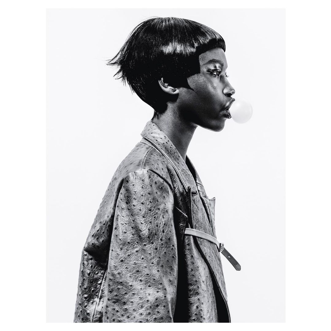 Black and white portrait of an African American woman blowing a bubble with bubble gum, wearing a textured coat, against a white background.