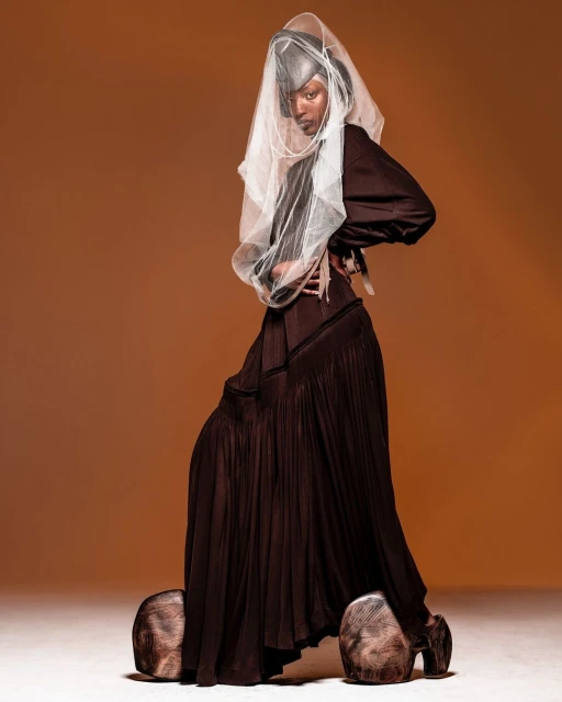 Woman in brown dress with oversized wooden shoes, posing confidently against a solid brown backdrop in a studio setting with soft lighting.