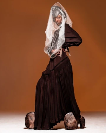 Woman in brown dress with oversized wooden shoes, posing confidently against a solid brown backdrop in a studio setting with soft lighting.