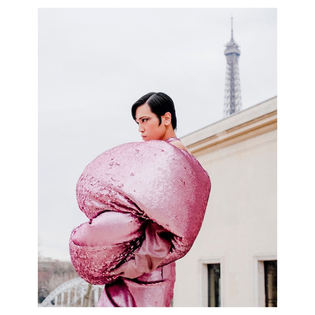 Person in a voluminous pink sequin dress stands in front of the Eiffel Tower. Fashion editorial portrait with an urban backdrop.