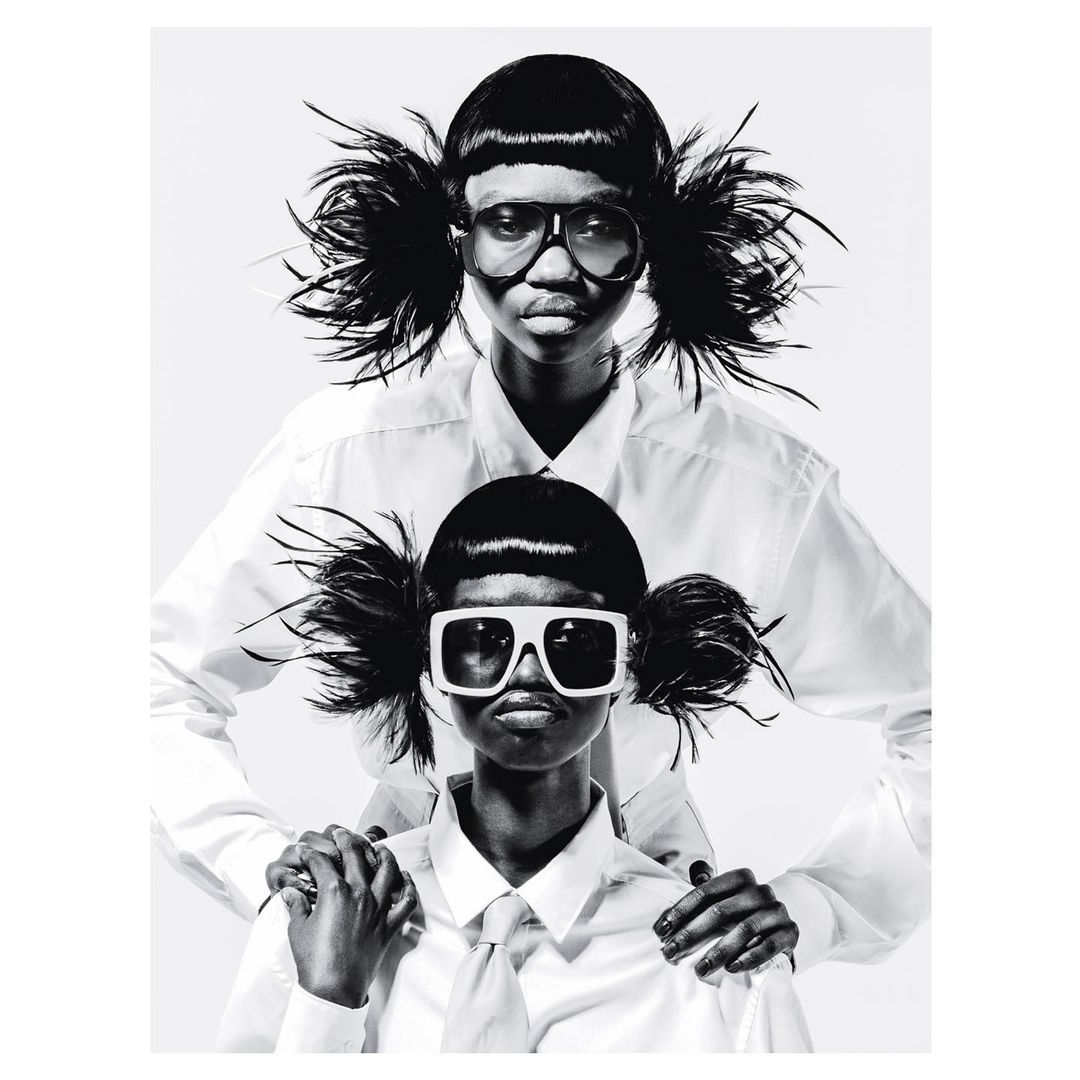 Striking black and white fashion portrait of two dark-skinned adult females with matching hairstyles, white shirts, and sunglasses in a photo studio.