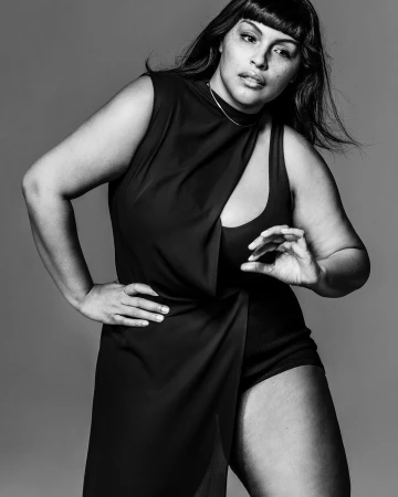 Elegant plus-size woman in a black dress poses in a studio portrait. Stylish one-piece with cutouts and draped skirt. Fashion and beauty.