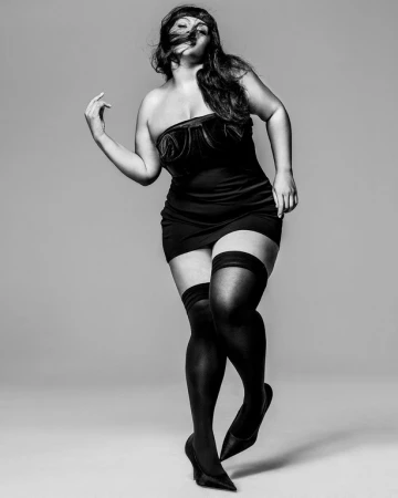 Monochrome fashion photograph of a woman wearing a strapless black dress and thigh-high stockings against a grey background.