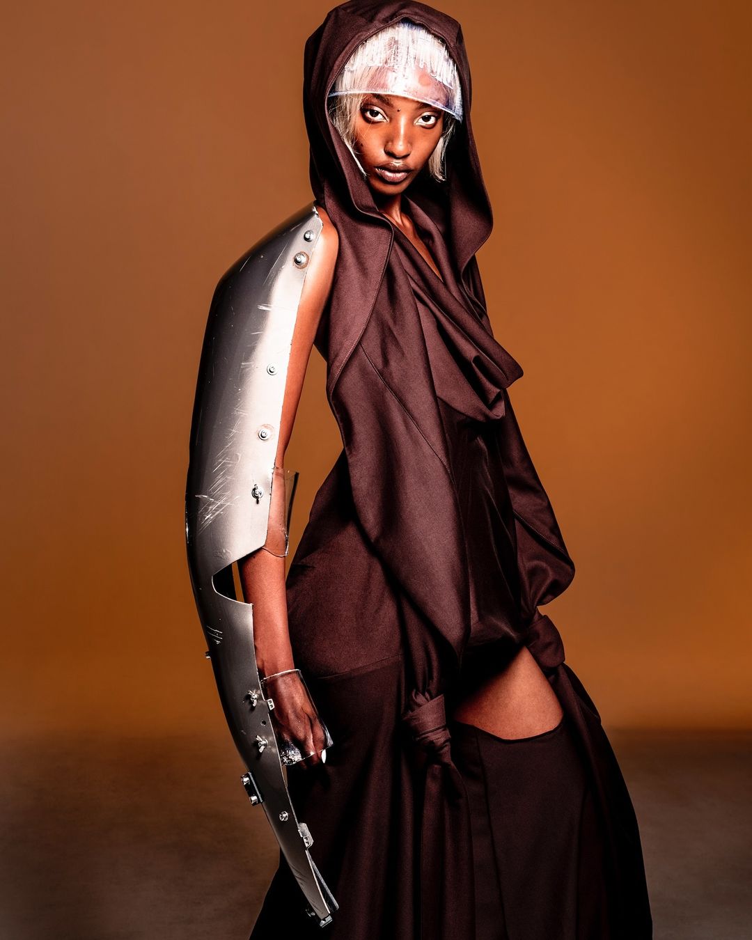 Woman in brown hooded gown poses with a futuristic silver arm brace against a warm brown backdrop in a studio shot. Fashion-forward.