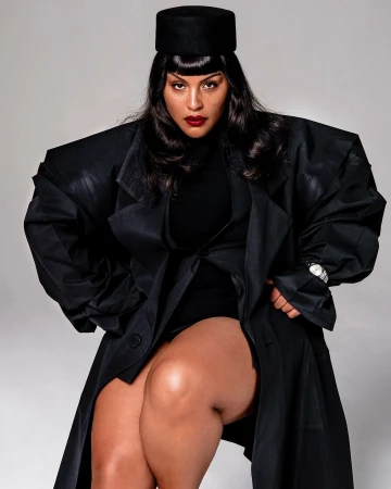 Woman in an oversized black coat with structured shoulders and a pillbox hat photographed by Cruz Valdez against a grey backdrop.