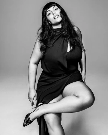 Black and white portrait of a woman in a black dress with high heels against a grey seamless studio background.