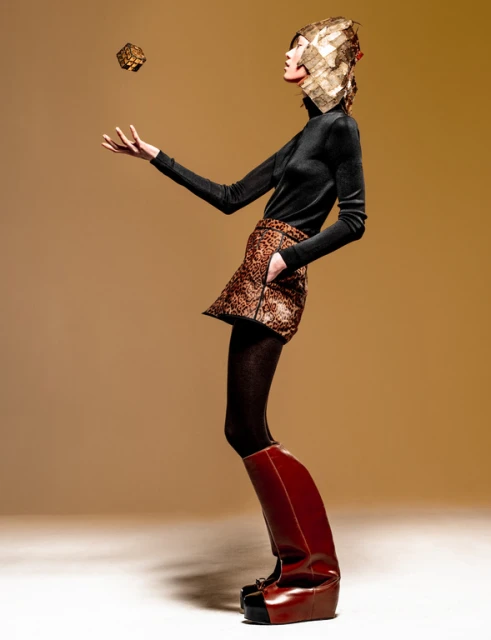 Fashionable woman in black top, patterned skirt, and unique boots poses with geometric object in studio with soft, warm lighting.