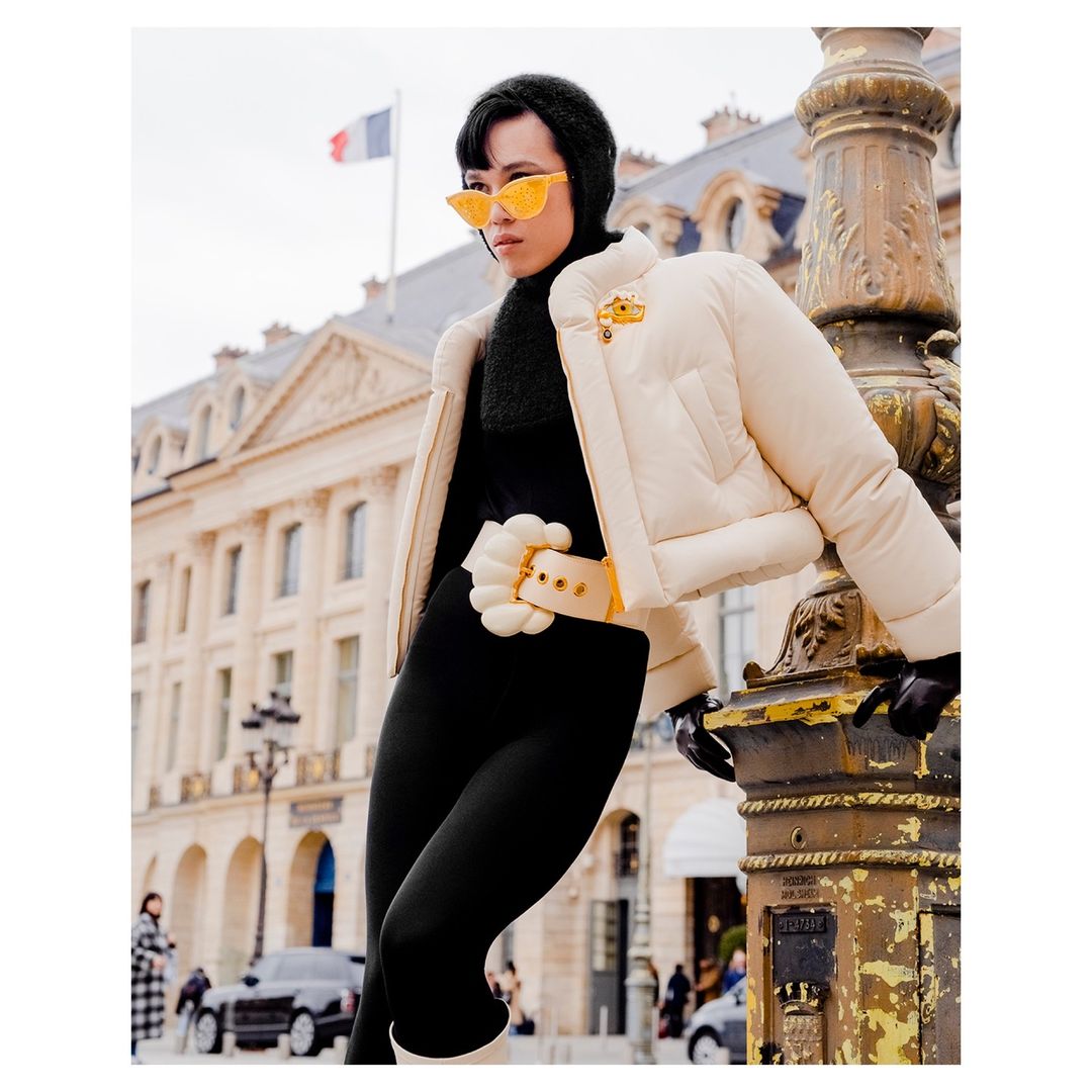 Woman in a cream puffer jacket, black leggings, and yellow glasses stands next to a vintage street lamp post in Paris, France.