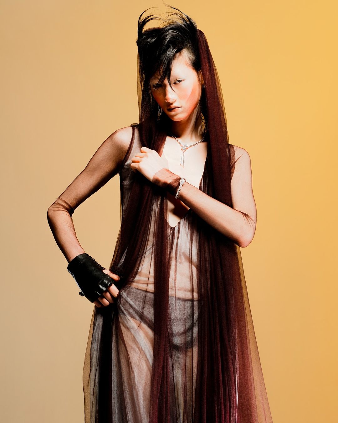 A dramatic fashion shot of an Asian woman with stylized hair and makeup posing in a sheer dress on a tan background.