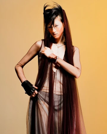 A dramatic fashion shot of an Asian woman with stylized hair and makeup posing in a sheer dress on a tan background.