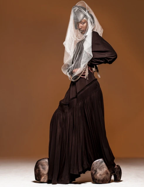 Model wears brown dress and avant-garde head covering with white net, posing in studio against a brown background in a fashion shot.