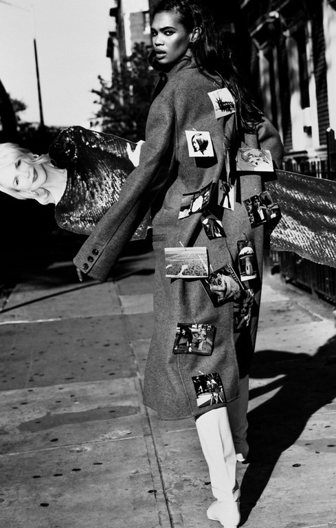 Woman in oversized grey coat decorated with photos, paired with white boots, holds sequined garment on city sidewalk.