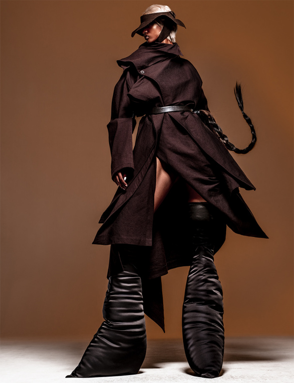 Model in a dark oversized belted coat, wide-brimmed hat, and exaggerated knee-high boots against a warm solid background.