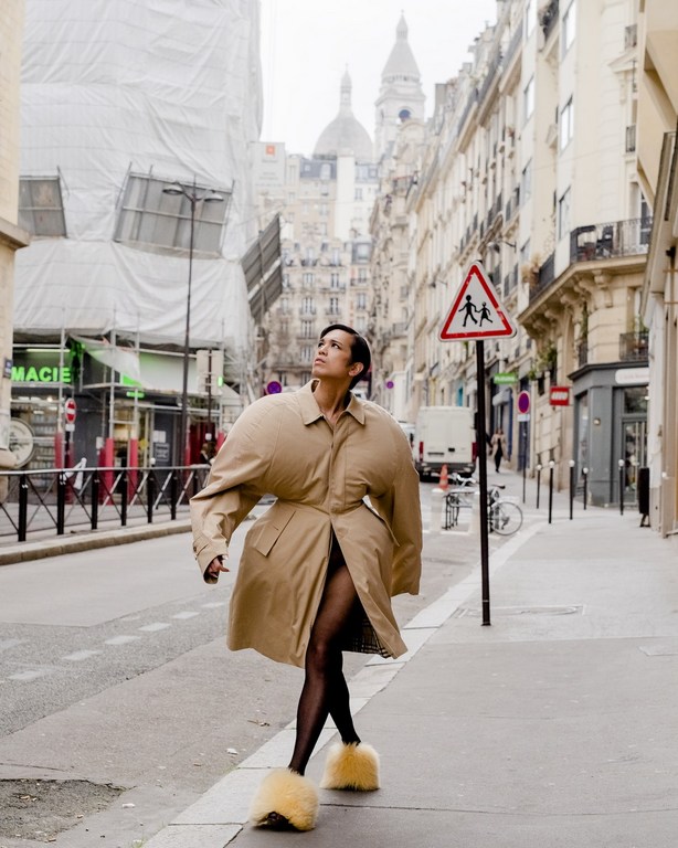 Fashionable woman in oversized beige trench coat walks on Paris street, wearing black tights and furry slides, in urban fashion editorial.