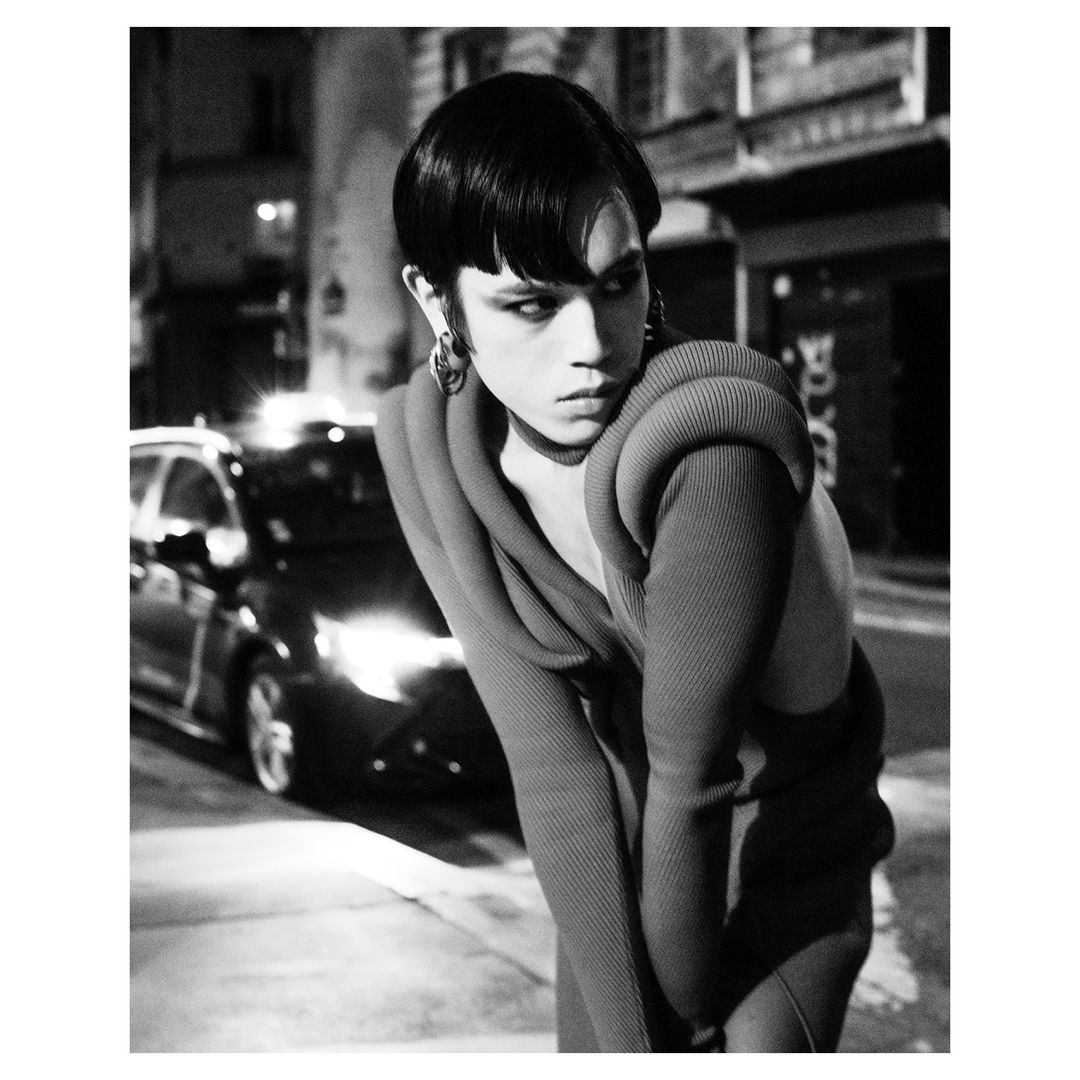 Black and white studio shot of a person with a short, dark hairstyle wearing a sweater with exaggerated shoulder details on a city street.