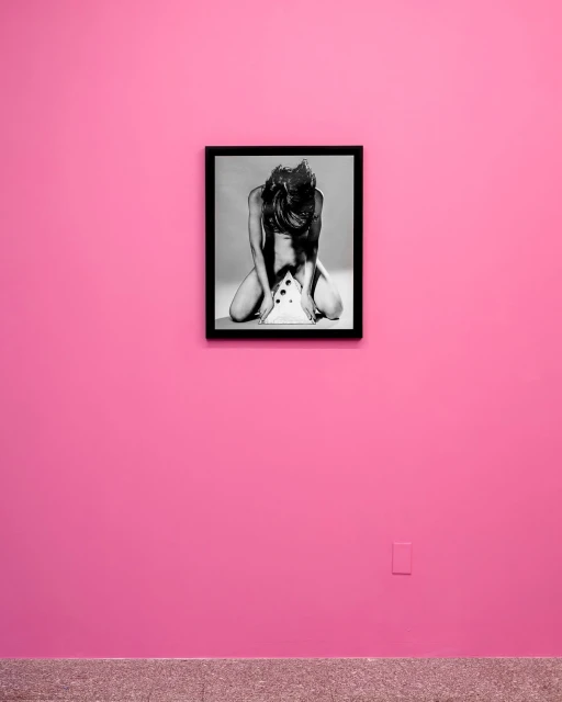 Monochrome photograph of a woman kneeling over a white object, framed and mounted on a pink wall above a grey baseboard.