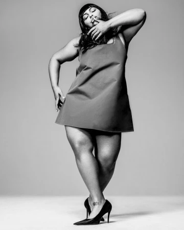 Black and white portrait of a woman in a short dress and heels posing against a seamless grey backdrop.