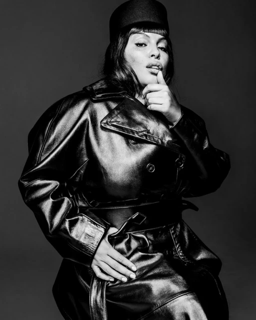Black and white portrait of a woman in a leather trench coat and hat, posing against a grey backdrop in a photo studio.