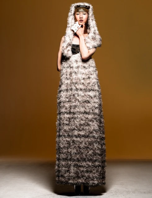 Woman posing in a shaggy dress and hood against a yellow background in a studio setting, holding playing cards in her hand.