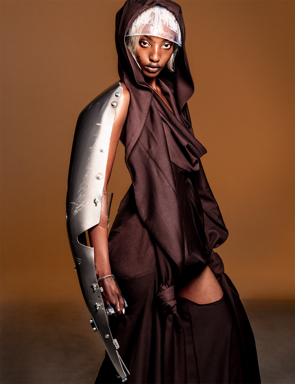 Fashionable adult woman in hood and gown wearing an arm-length metal gauntlet poses in a studio with a neutral background.