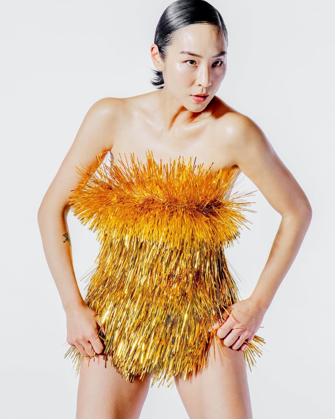 Asian woman poses in a short, strapless gold metallic fringe dress against a plain white backdrop in a fashion-style studio portrait.