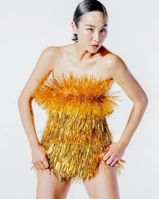 Asian woman poses in a short, strapless gold metallic fringe dress against a plain white backdrop in a fashion-style studio portrait.